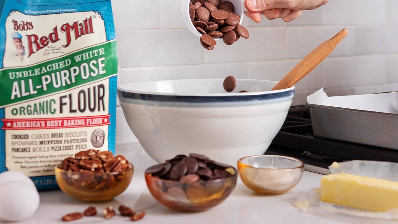 Chocolat-e baking chips are being poured into a large white mixing bow. A package of Bob's Red Mill All-Purpose Organic Flour, eggs, pecans, and butter surround the bowl.