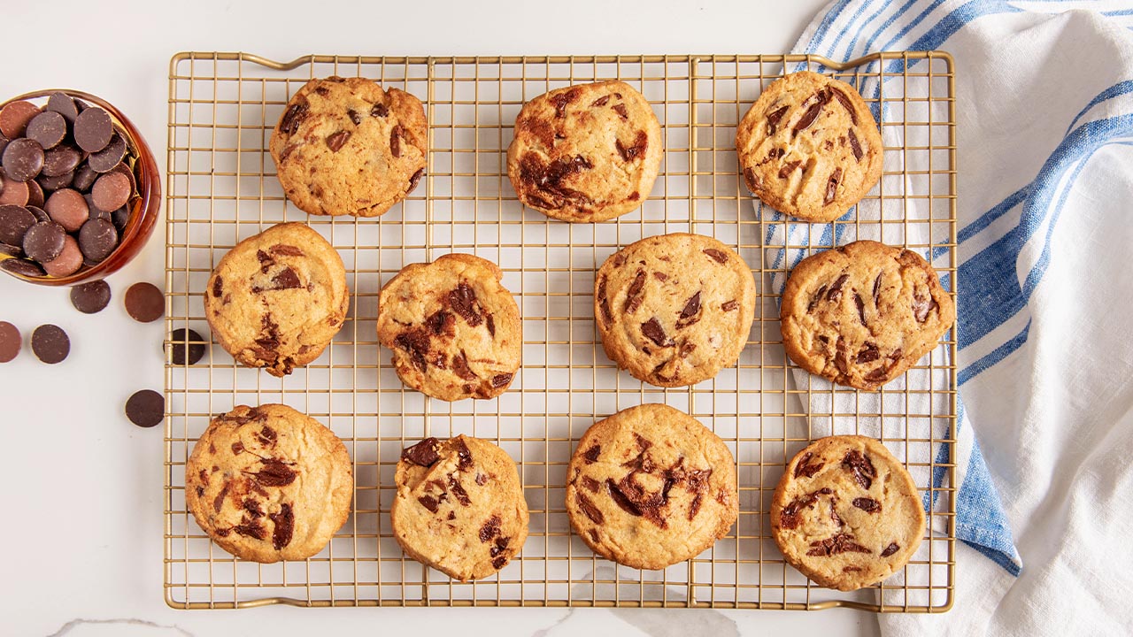 Freshly baked chocolate chip cookies are on a cooling rack. A bowl of baking chips with both dark chocolate and dark-milk chocolate, is to the left. 