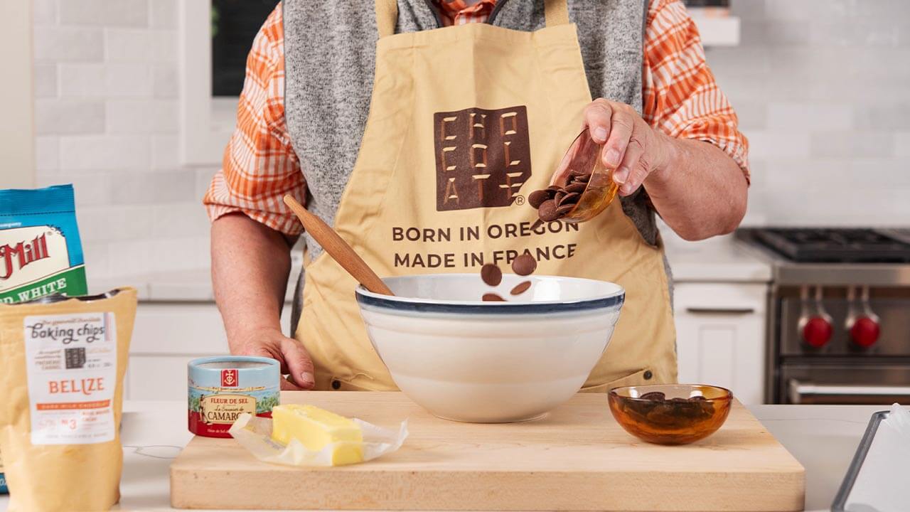 Chocolat-e baking chips are being poured into a large white mixing bowl. A cup of additional baking chips, butter, flour, and other ingredients are also pictured.