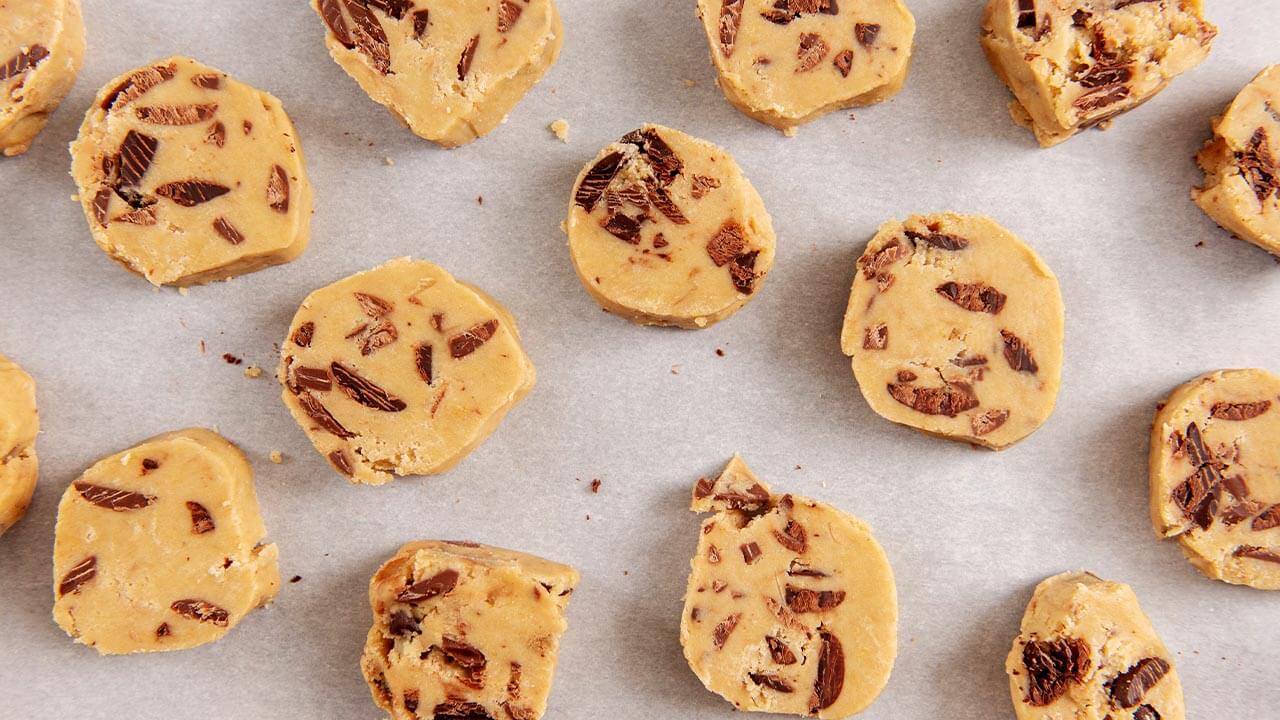 Slices of chocolate chip cookie dough are placed on parchment paper. 