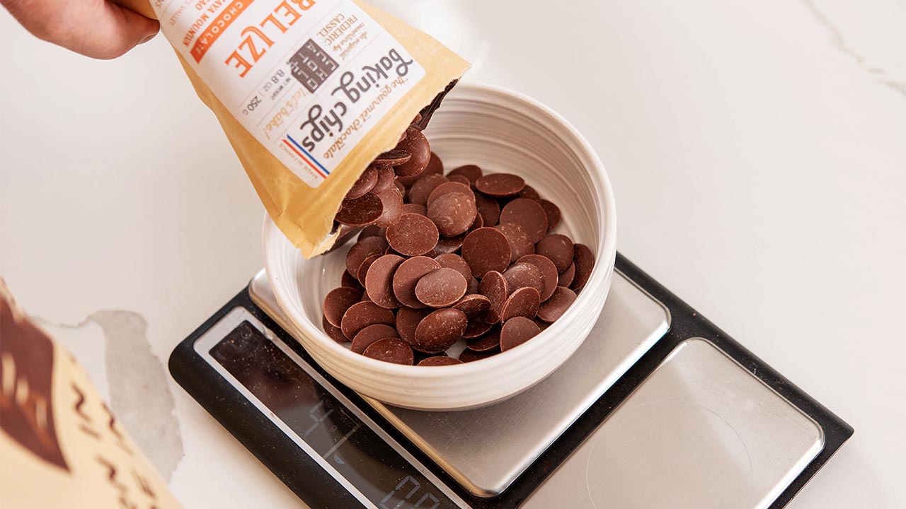 Dark-milk chocolate baking chips from Belize are being poured from the Chocolat-e packaging into a small white bowl on a food scale.