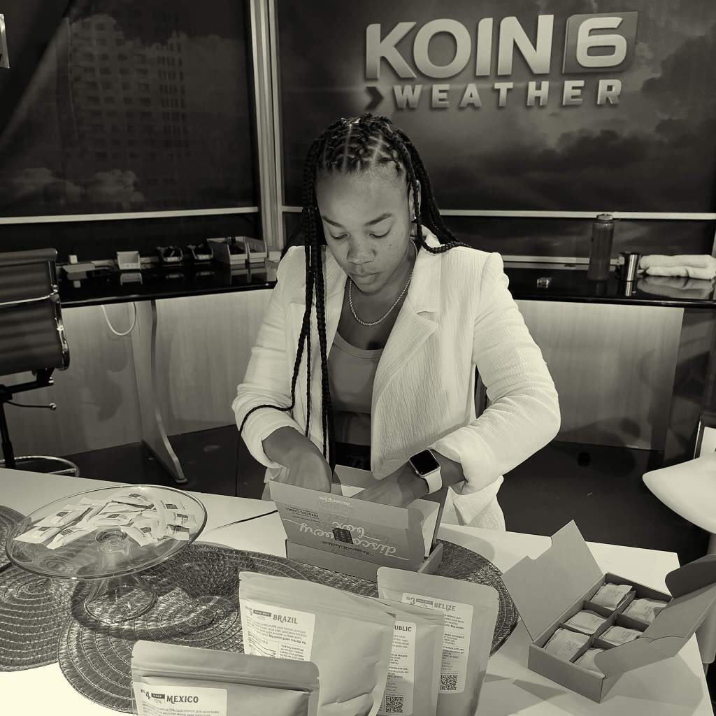 A reporter at KOIN 6 news is opening a Chocolat-e discovery box on a table filled with other Chocolat-e items.