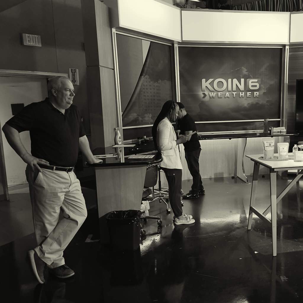Behind the sceens at KOIN 6 weather desk. Three people are standing and one is on their cell phone.