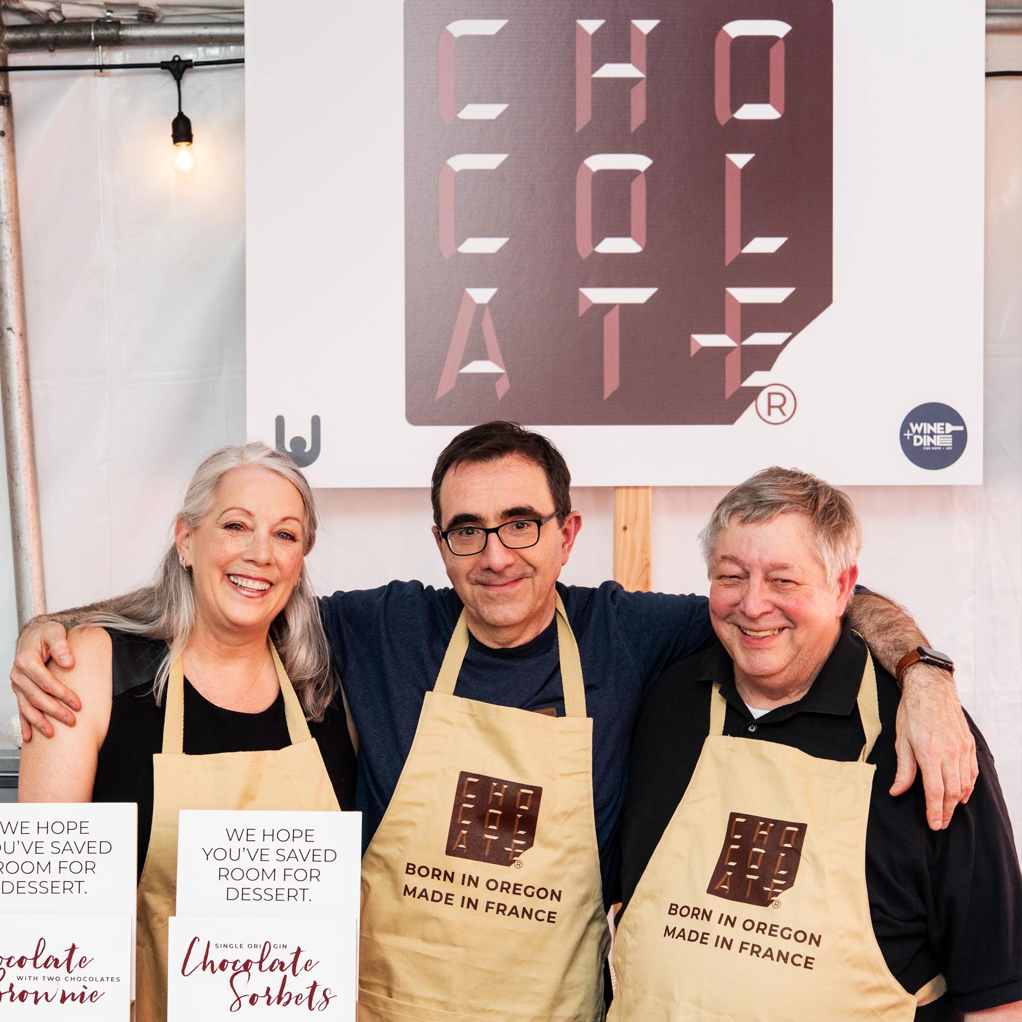 Jeanyves and Norm are joined by another person, all are wearing matching Chocolat-e aprons, and smiling.